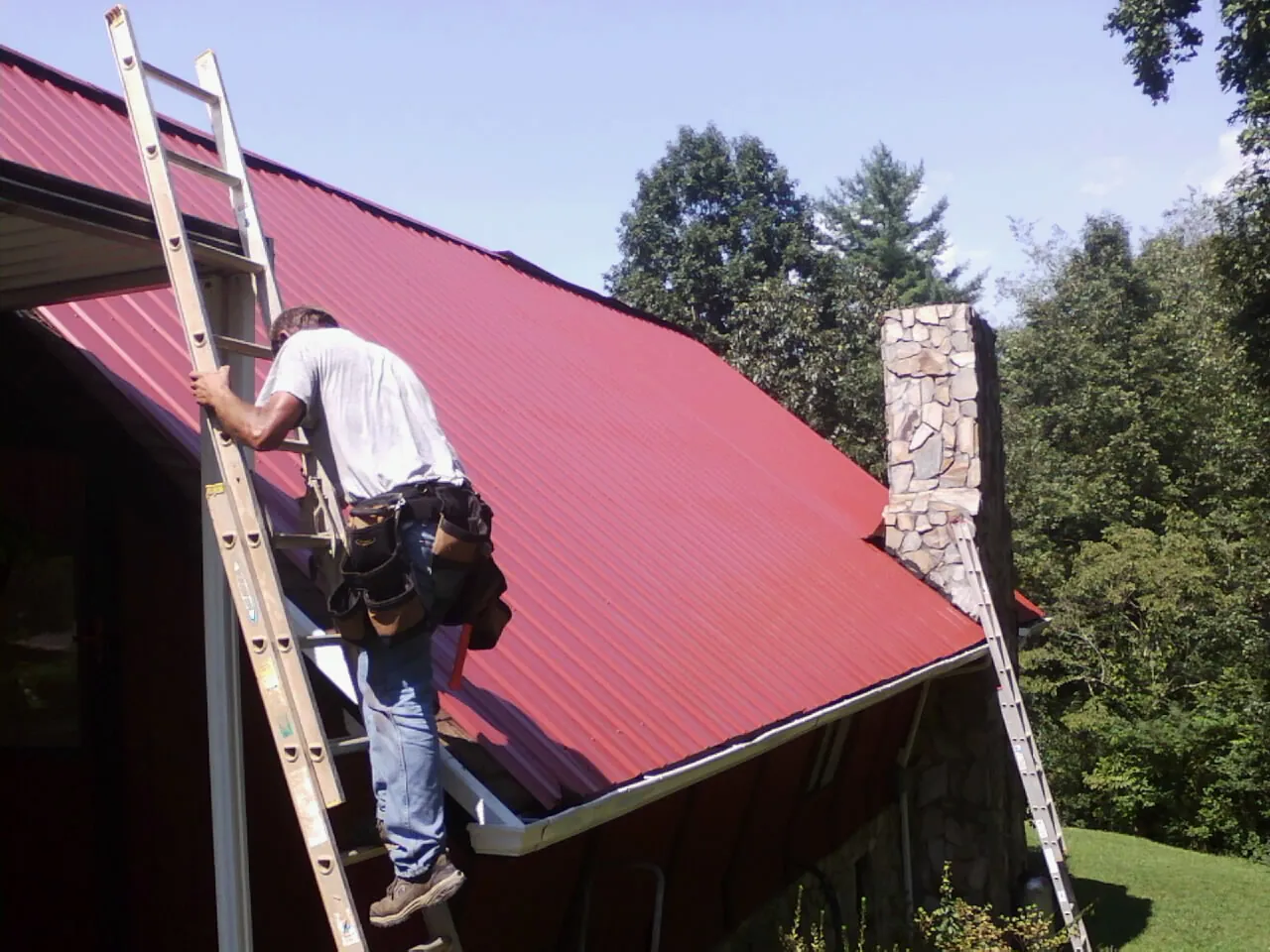 Roofer on a ladder beside a red metal roof and stone chimney — Soffit & Fascia Repair by Roofing Brothers in Lodi